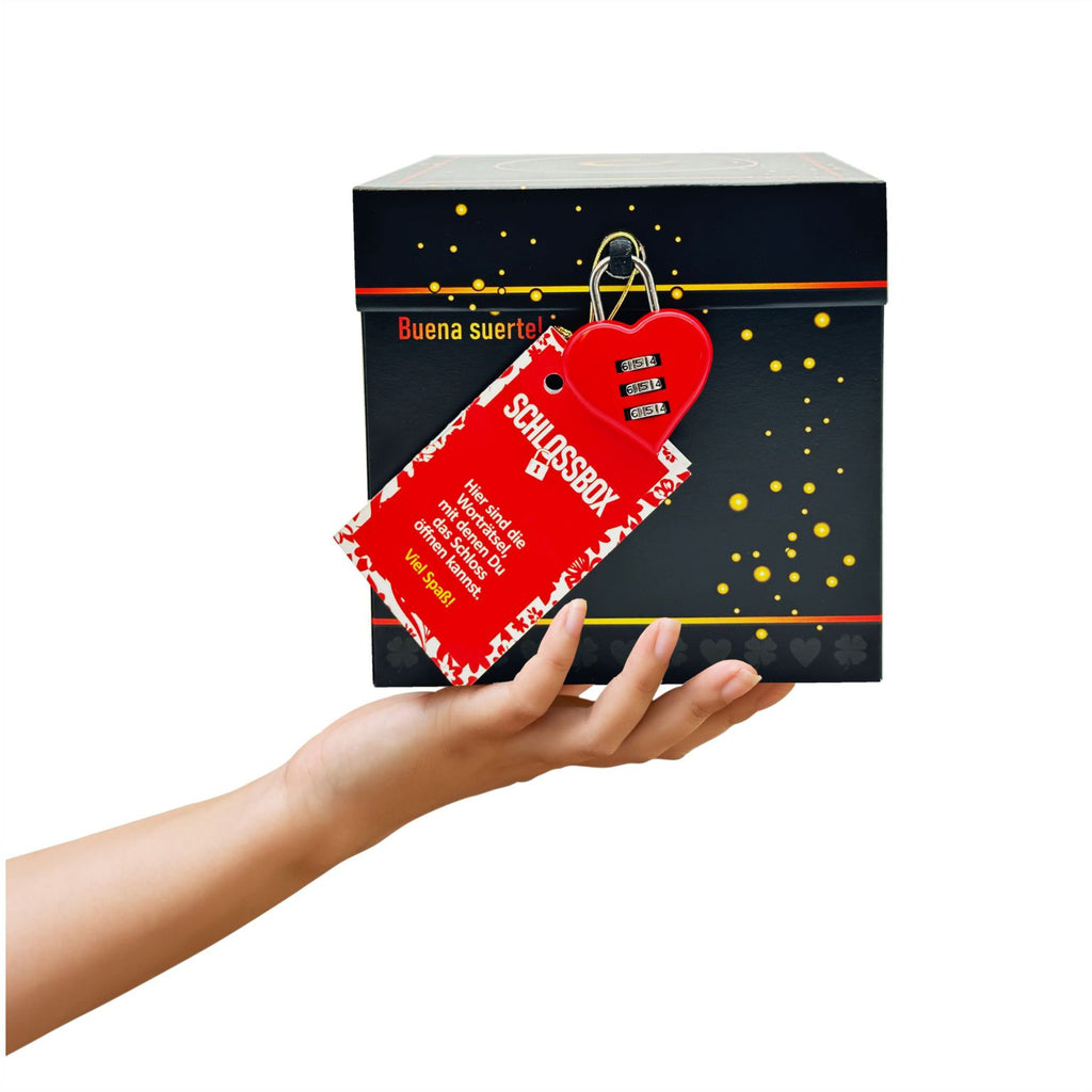 Rätselbox Glück in Schwarz mit rotem Herz-Zahlenschloss und Rätselheft-Anhänger, gehalten in einer Hand vor weißem Hintergrund. Aufschrift vorne an der Box: Buena suerte!