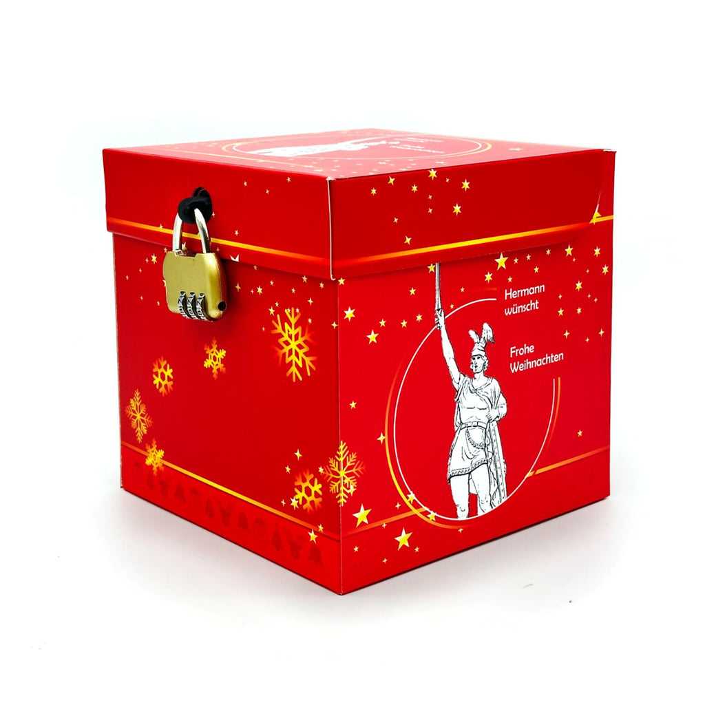 Geschlossene Rätselbox Weihnachten Hermannsdenkmal in Rot mit goldenen Stern- und Schneeflockenmotiven sowie angebrachtem goldenem Zahlenschloss. Auf der Seite eine Illustration des Hermannsdenkmals. Text: Hermann wünscht Frohe Weihnachten