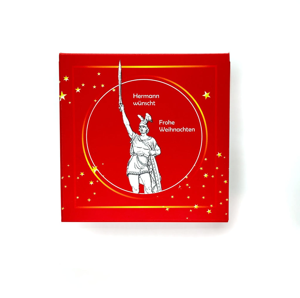 Deckel der Rätselbox Weihnachten Hermannsdenkmal in Rot mit goldenen Sternen und Motiv des Hermannsdenkmals mit Schriftzug ‚Hermann wünscht Frohe Weihnachten‘.
