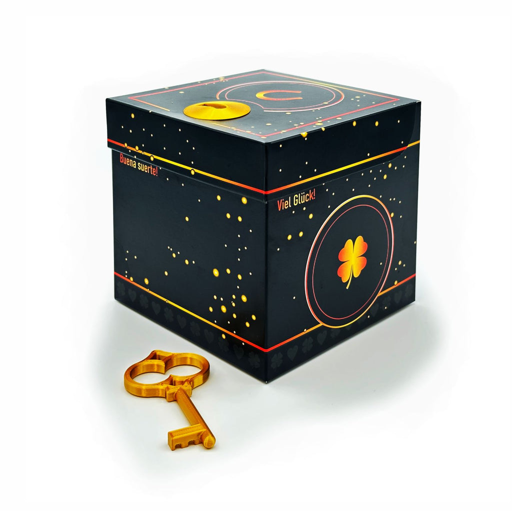 Geschlossene Schlossbox Glück in Schwarz mit goldenen Sprenkeln und Kleeblatt-Motiv, seitlich Schriftzüge ‚Viel Glück‘ und ‚Buena suerte‘, goldener Schlüssel liegt vor der Box