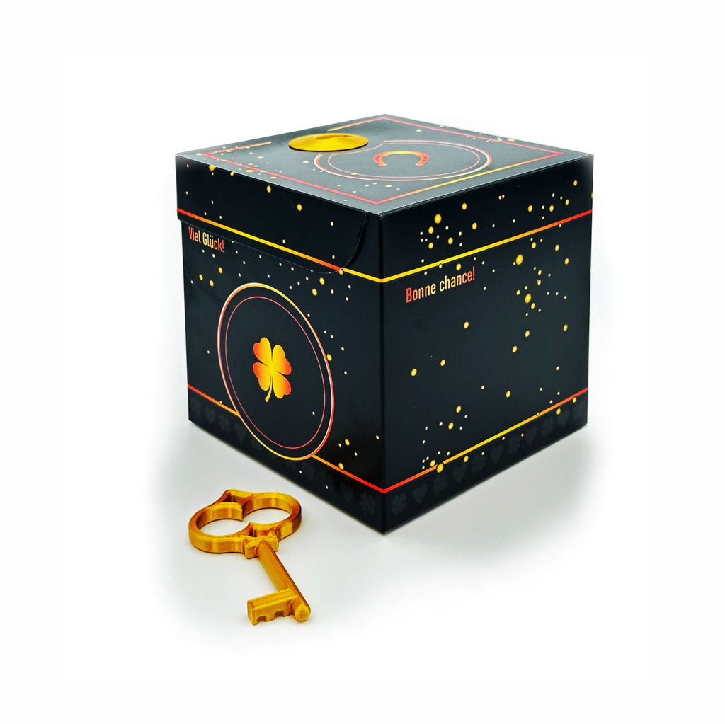 Geschlossene Schlossbox Glück in Schwarz mit goldenen Sprenkeln und Kleeblatt-Motiv, seitlich Schriftzüge ‚Viel Glück‘ und ‚Bonne chance‘, goldener Schlüssel liegt vor der Box.