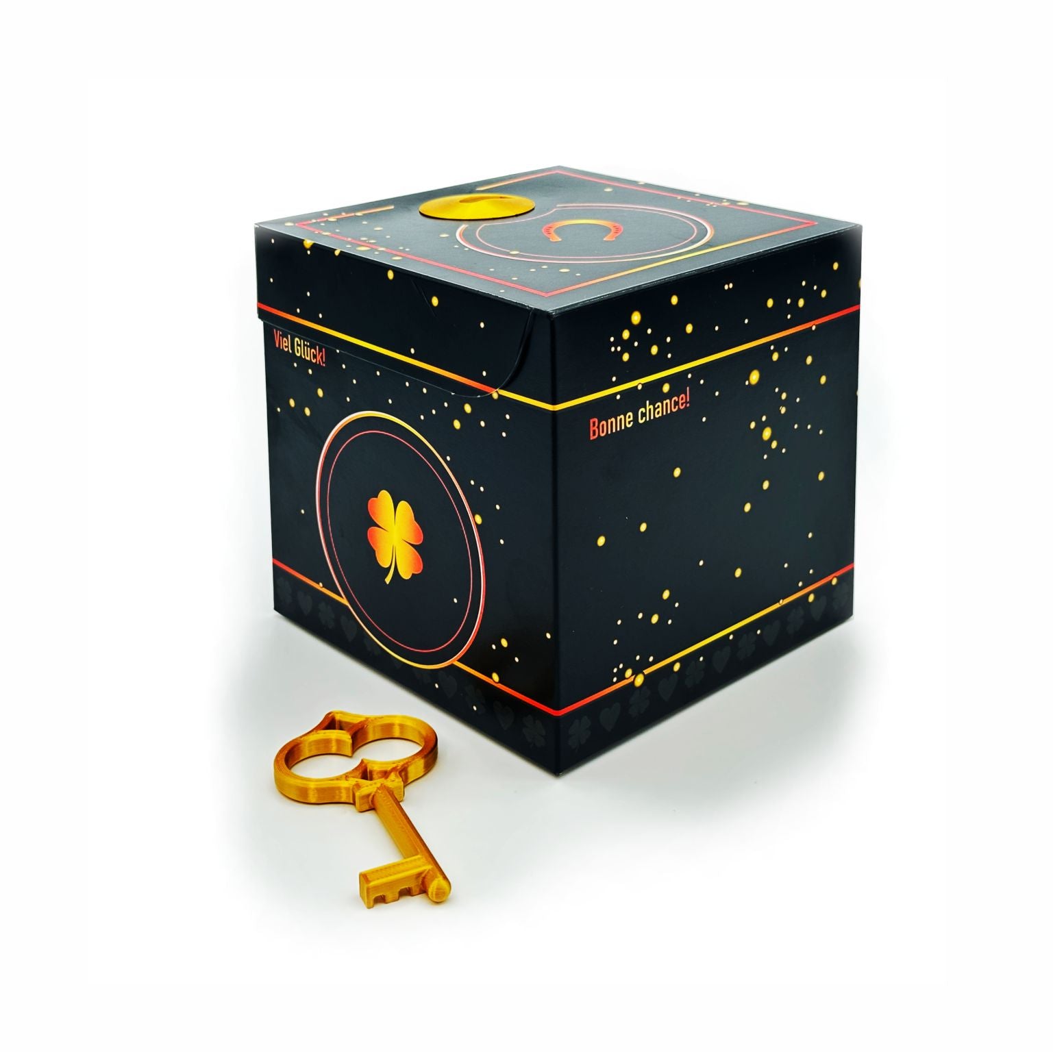 Geschlossene Schlossbox Glück in Schwarz mit goldenen Sprenkeln und Kleeblatt-Motiv, seitlich Schriftzüge ‚Viel Glück‘ und ‚Bonne chance‘, goldener Schlüssel liegt vor der Box.